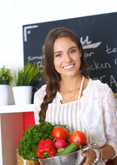 Wall Mural - Smiling young woman holding vegetables standing in kitchen