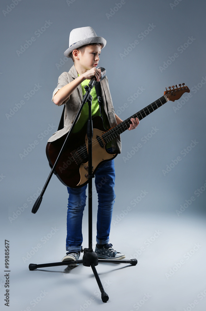 Little boy playing guitar and singing with microphone on grey background