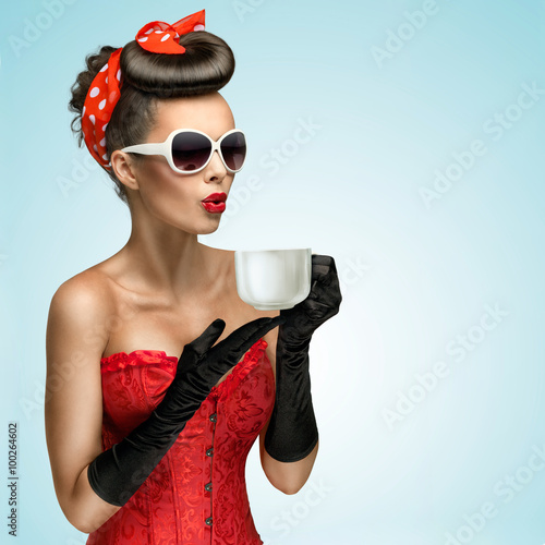 Naklejka na meble Hot beverage / Three-quarter portrait of glamourous pinup girl wearing vintage gloves and red ribbon in her hair, holding a cup of hot coffee or tea and cooling it.