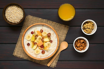 Wall Mural - Oatmeal porridge with grape, apple and banana in wooden bowl, walnut, almond, raw oatmeal, fresh juice and wooden spoon on the side, photographed overhead on dark wood with natural light