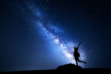 Wall Mural - Milky Way. Night sky with stars and silhouette of a happy woman with backpack and raised up arms. Space background