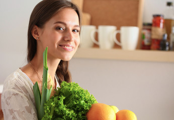 Wall Mural - Smiling young woman holding vegetables standing in kitchen