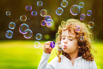 Curly little girl blowing soap bubbles in summer park.