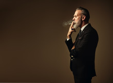 Portrait Of Smoking Gentleman Wearing Trendy Suit And Stands Against The Empty Wall. Horizontal