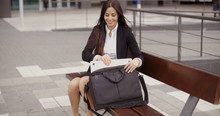 Attractive Stylish Young Businesswoman Sitting On A Wooden Urban Bench Placing Her Laptop In A Bag With A Smile