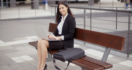 Wall Mural - Cute business woman on bench in front of office building looking over while working on laptop computer