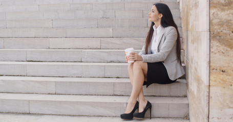 Wall Mural - Grinning brunette professional woman sitting on outdoor stairs while drinking coffee