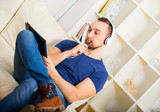 Fototapeta  - Handsome young man at home writing on notebook, sitting on couch, listen to music