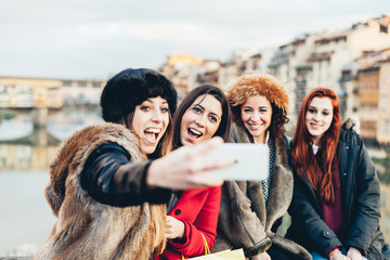 Four female friends taking a selfie on a bridge in Florence, Tuscany. Behind them the beautiful Ponte Vecchio. The women are dressed in winter jackets and hats against the cold of winter