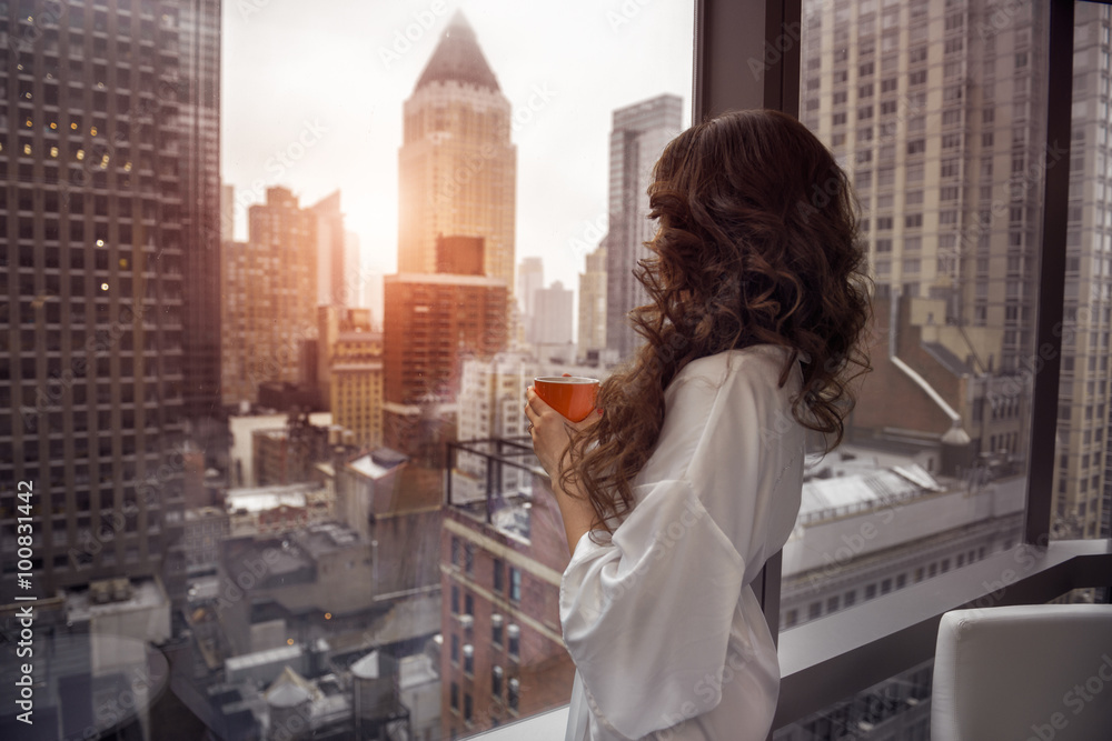 Beautiful woman holding coffee cup and looking to the window in luxury Manhattan penthouse apartments. Good morning after wake up. - obrazy, fototapety, plakaty 