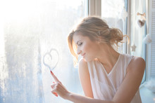  Beautiful Woman With Fresh Daily Makeup And Romantic Wavy Hairstyle, Sitting At The Windowsill, Draws On Glass