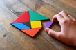 man's hand holding a missing piece in a square tangram puzzle, over wooden table.
