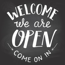 Welcome We Are Open. A Welcome Sign For Cafes Or Shop Visitors On Blackboard Background With Chalk. Hand Lettering