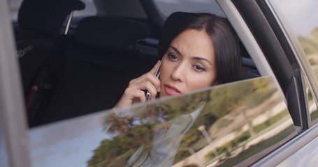 Wall Mural - Serious young woman in business attire on cell phone while sitting in rear seat of limousine looking outside from open window