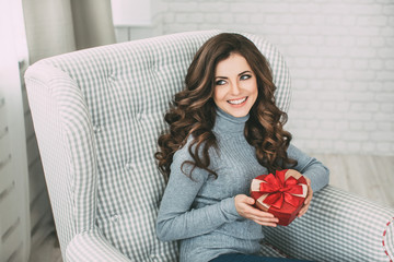 Wall Mural - Young beautiful smiling brunette with long curly hair sitting in a chair and holding a gift.