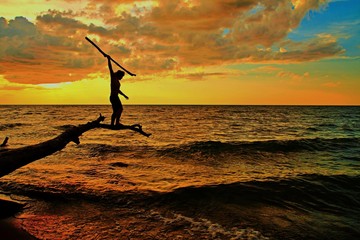 The Hunt. Silhouette of a male balanced on a log over the water preparing to spear a fish.