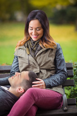 Wall Mural - Happy young woman sitting on a bench in a park with her boyfriend lying on her lap