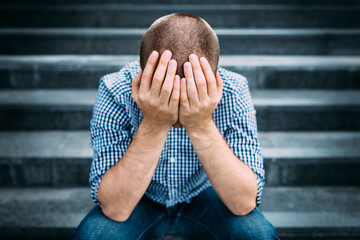 Outdoor portrait of sad young man covering his face with hands sitting on stairs. Selective focus on hands. Sadness, despair, tragedy concept