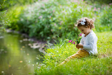 Sweet, Happy Little Girl Sitting On A Grass In A Park At A Spring Stream With Flower In Hand. Laughing, Enjoying Fresh Air In Forrest. 
