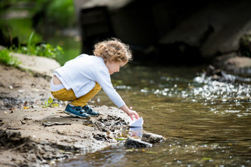 Wall Mural - Cute little girl runs a paper boat in the stream in the park. Stretching her hand and reaching the little ship