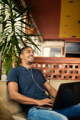 Wall Mural - Handsome black man sitting at cafe bar,listening music and typing on laptop. He is looking at computer and smiling.