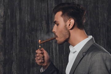 Wall Mural - Handome brutal man in suit on the grey background lighting a cig