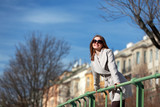 Fototapeta Miasto - Beautiful blonde woman with coat and sunglasses leaning on a handrail and enjoying the sun