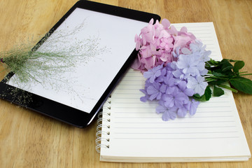 Digital tablet and notebook with bouquet of flowers on wooden table.