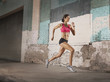 © Mint Images - A woman running along an urban street past buildings with peeling paint and a metal shutter.