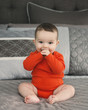 Portrait of cute adorable Caucasian smiling laughing baby boy girl with black brown eyes in orange red onesie shirt sitting on bed looking directly in camera, natural window light, lifestyle