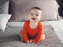 Portrait Of Cute Adorable Caucasian Smiling Laughing Baby Boy Girl With Black Brown Eyes In Orange Red Onesie Shirt Sitting On Bed Looking Away From Camera, Natural Window Light, Lifestyle