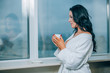 © satyrenko - Getting warm with fresh coffee. Beautiful young woman in white bathrobe drinking coffee and looking through a window