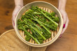 Girl holding bamboo pan with steamed broccoli rabe. Broccoli is prepared and ready to eat.
