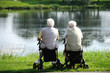 Zwei Rentner sitzen auf Rollatoren und sehen auf einen See.