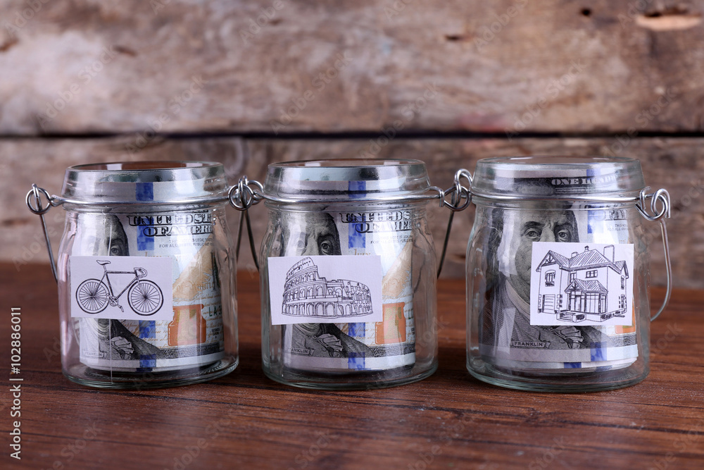 Rolls of dollar banknotes for different needs in glass jars on wooden table