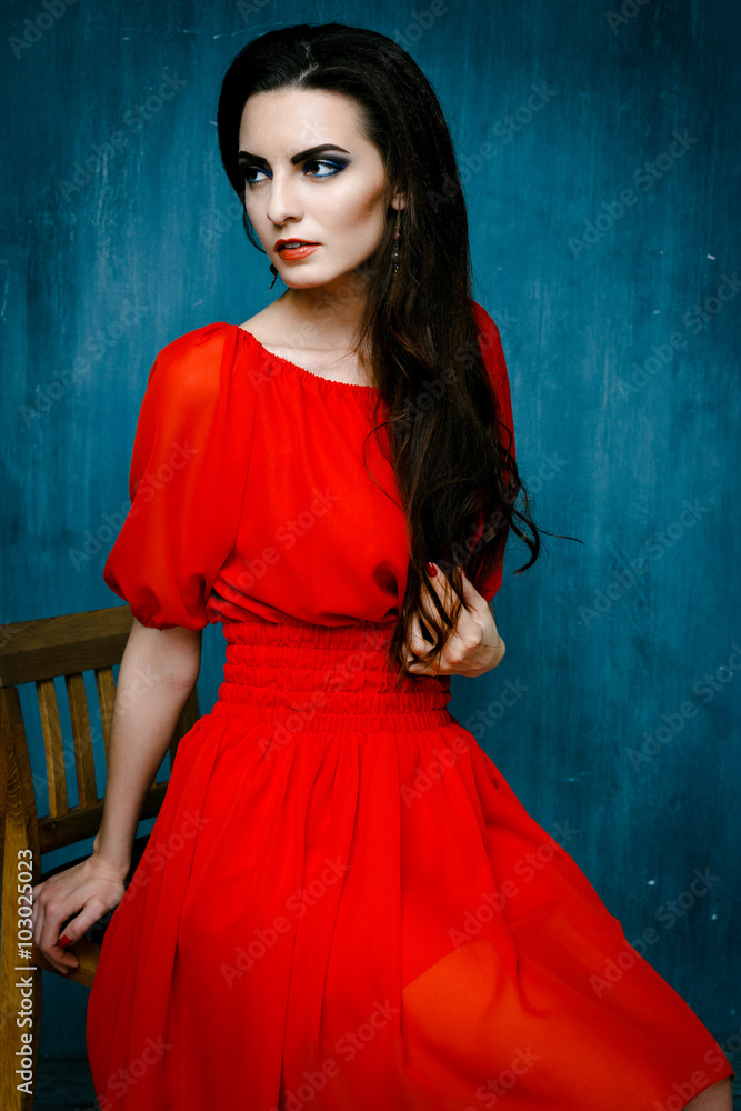 Beautiful elegant female model posing in red dress with red lips dark make up on blue background. Beauty Woman. Beautiful Brunette Luxury Girl. Healthy Long Black Hair and Holiday Makeup. Stock Photo | Beautiful elegant female model posing in red dress with red lips dark make up on blue background. Beauty Woman. Beautiful Brunette Luxury Girl. Healthy Long Black Hair and Holiday Makeup. Stock Photo |