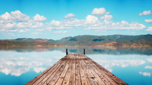 Perspective View Of A Wooden Pier In A Completely Calm Lake With Reflections Of The Sky