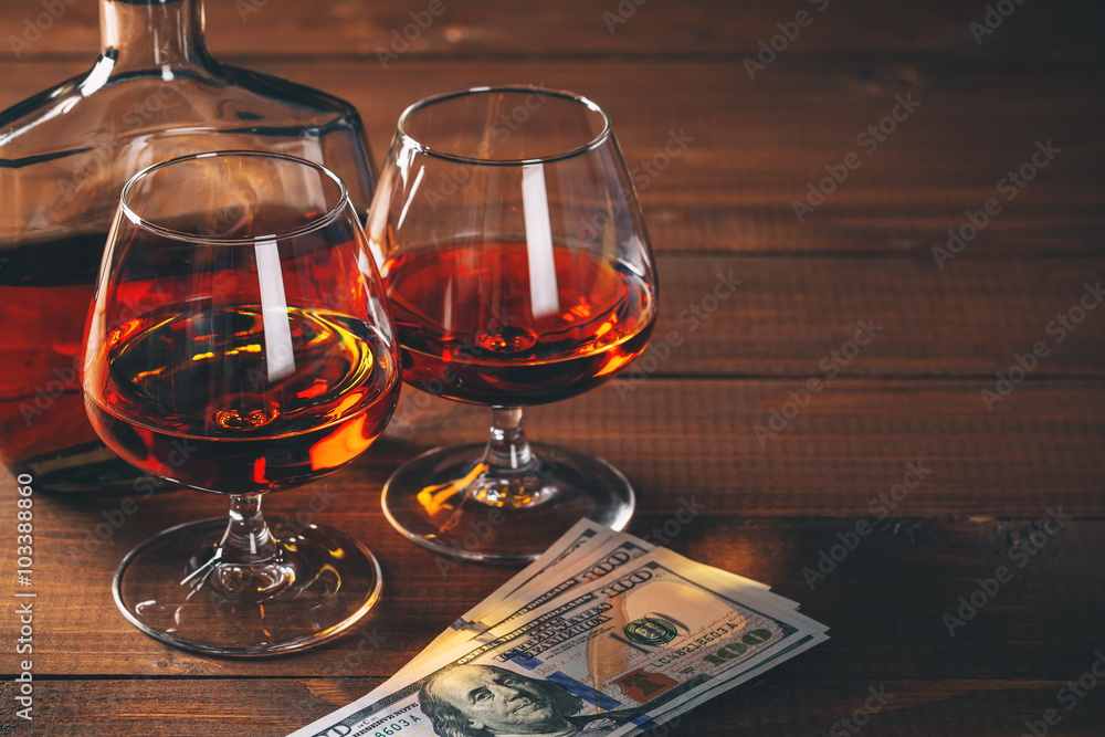 Two glasses of cognac and bottle, with wad of money on the wooden table.