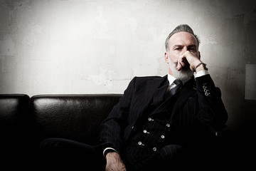 Portrait of adult gentleman wearing trendy suit and sitting modern studio on leather sofa against the empty concrete wall. Horizontal