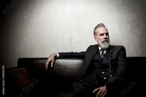 Fotografia Portrait of adult businessman wearing trendy suit and sitting modern studio on leather sofa against the empty concrete wall