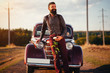 © endrews21 - brutal bearded man with a mustache in a shirt, pants with suspenders stands leaning on the hood of a retro car holding a white rose and looking at the sunset