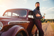 © endrews21 - brutal bearded man with a mustache in a shirt, pants with suspenders stands leaning on the hood of a retro car holding a white rose and looking at the sunset