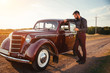 © endrews21 - brutal bearded man with a mustache in a shirt, pants with suspenders stands leaning on the hood of a retro car holding a white rose and looking at the sunset
