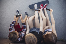 Three Teenagers Sitting Outdoors With Smartphones And Skateboard