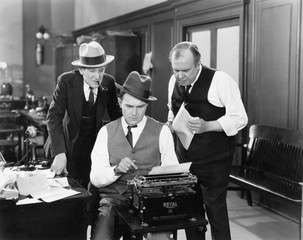 three men in an office hunched over a typewriter