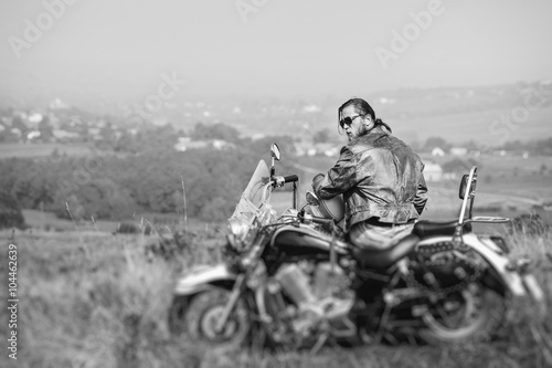 Tapeta ścienna Brutalny rowerzysta z brodą na sobie skórzaną kurtkę i okulary przeciwsłoneczne, siedząc na motocyklu w słoneczny dzień, trzymając hełm. 