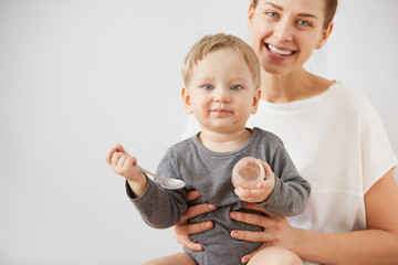 Young attractive mother with her one years old little son dressed in pajamas.  Boy eating a fruit smoothie himself in the bedroom at the weekend together, warm and cozy scene. Selective focus.