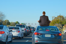 A Man Is Dealing With Intense Work Rush Hour Traffic Jam Stress By Getting Relief Doing Yoga On Top Of His Car In This Humorous Scene That Shows PEACE On The License Plate Of The Car He Is Sitting On.