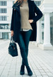 Woman in a sweater, black coat and pants holding a handbags whil
