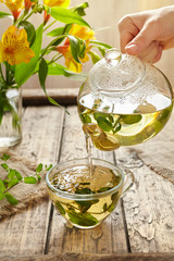 Wall Mural - Peppermint tea pouring in glass cup from teapot, yellow flowers on vintage wooden background. Healthy natural organic aromatherapy beverage.